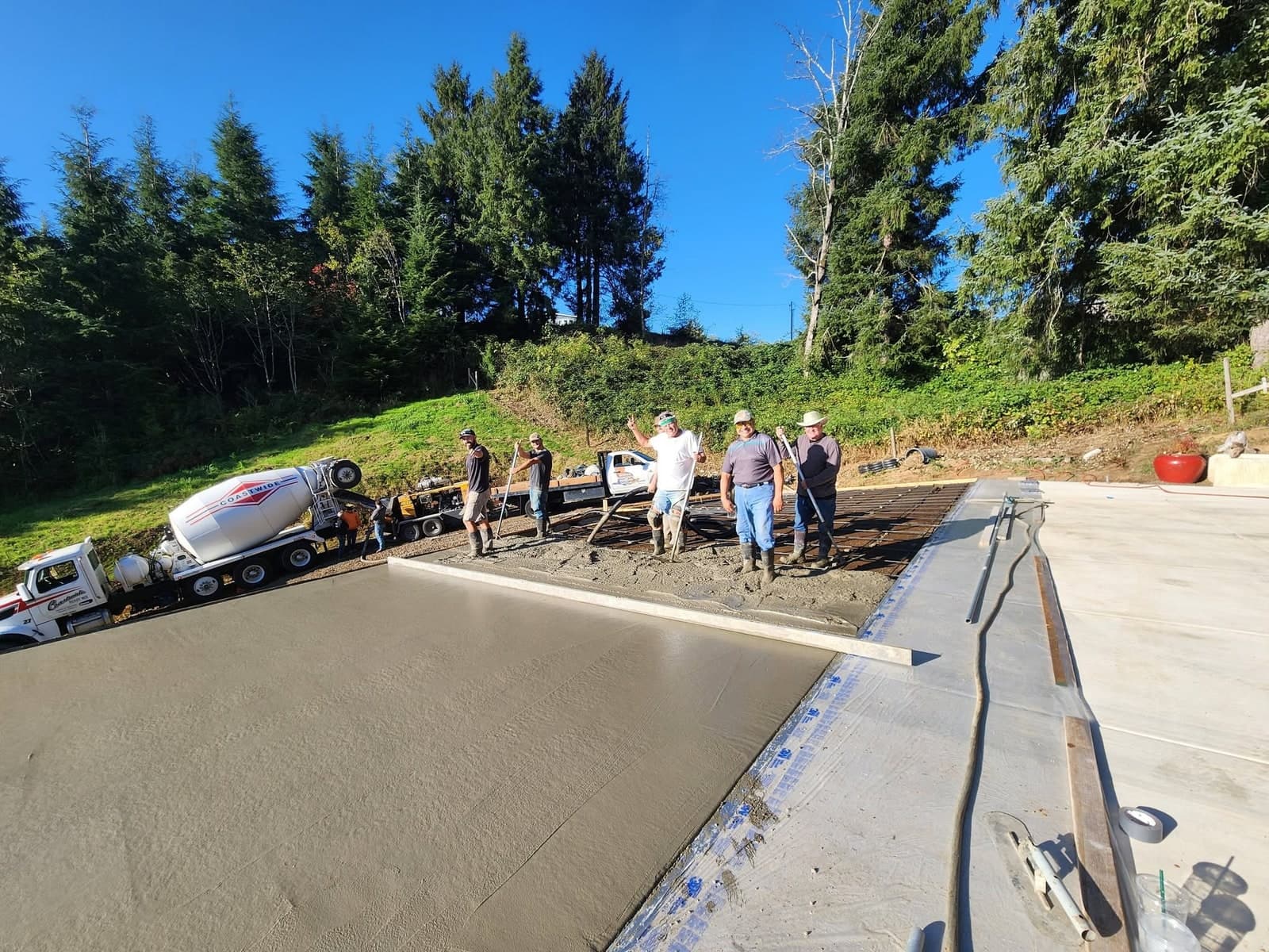 Five 5 Rivers crew standing along the far edge of a large freshly floated concrete slab, screeds and floats still in their hands and one of them with an arm raised toward the camera, a ready-mix truck parked up on the bank behind them and firs against a clear blue sky
