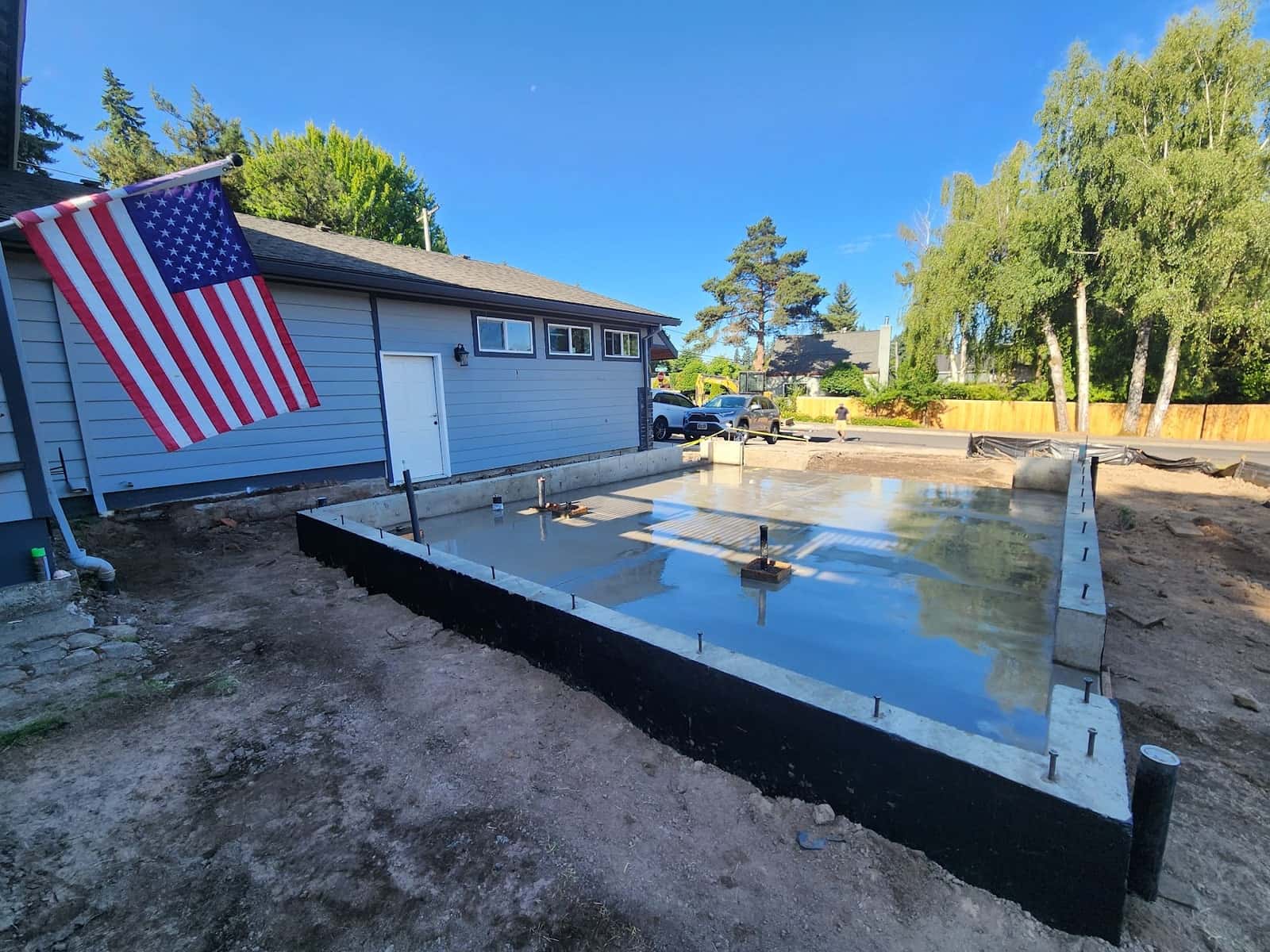 A finished concrete stem wall and slab, still wet and mirroring the sky, poured against a house with an American flag hanging from the garage wall